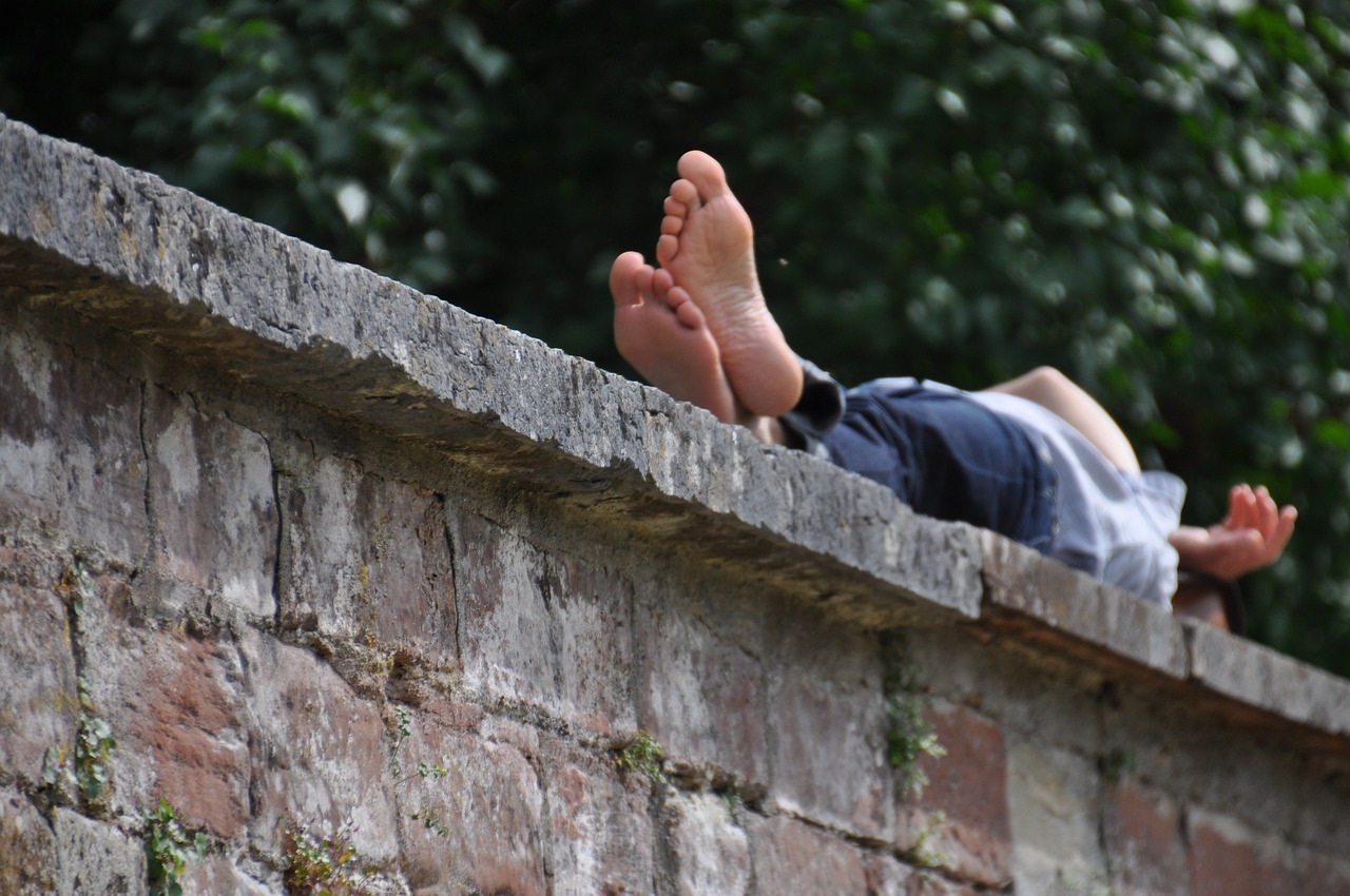 Person liegt entspannt barfuß auf einer alten Steinmauer, umgeben von grüner Natur. Die Füße sind im Vordergrund zu sehen, während der Oberkörper entspannt nach hinten gelehnt ist – ein Moment der Ruhe und Achtsamkeit in der Natur.